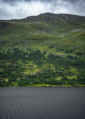 Fototapeta premium Holiday homes sit by a lake surrounded by green, beneath the steep mountain range. Storm clouds gather above the rocky outcrop, contrasting with the serene scene