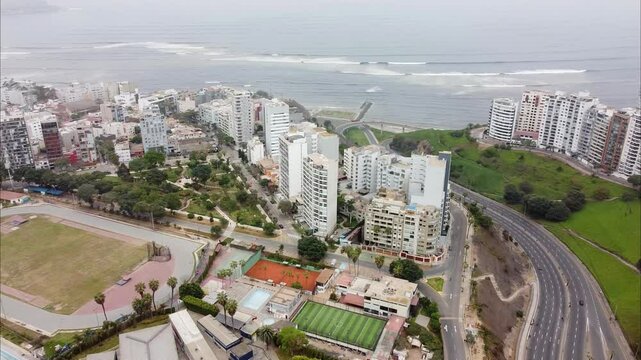 Aerial view of coastal city with high-rise buildings, parks, and scenic shoreline. Roads winding