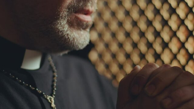 Mature priest holding hands together and praying with eyes closed in confessional booth. Close-up shot