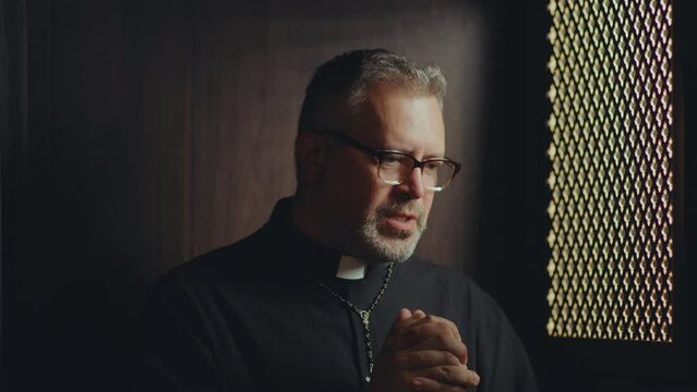 Mature Catholic priest wearing black cassock sitting inside of confessional booth, holding hands together and praying to God