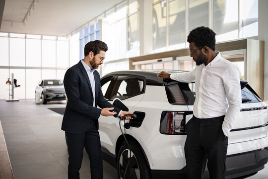 Dealer sells electric SUV to businessman in modern car dealership. Business negotiation about environmentally-friendly vehicle. Modern showroom conveys professional atmosphere. - Powered by Adobe