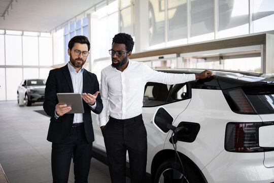 Car salesman using tablet to explain features of electric SUV to potential buyer in modern dealership. Business meeting focuses on environmentally friendly transportation and automotive technology