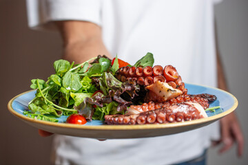 plate with grilled octopus and salad, held by man in white shirt