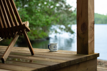 A coffee mug with the phrase "Life is better at the Cottage" rests beside a chair on the wooden porch of a boathouse, overlooking the serene blue waters of a lake in Muskoka, Ontario, Canada.