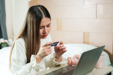 Asian woman working at home, examining credit card details, laptop and pink cup on table. Cozy bedroom setting, modern lifestyle, illustrating remote work and careful online shopping practices.