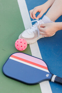 Hands tying shoe next to pickleball paddle and ball on court.