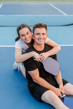 Couple smiling on a pickleball court, sitting, hugging with paddles.