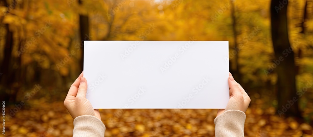Poster hands holding a blank white sheet with autumn leaves in the background, perfect for adding copy spac