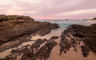 Fototapeta premium Sedimentary rocks at Currumbin estuary leading the eye across the bay to the tourist destination of Coolangatta which is under an overcast sky at sunset on the Gold Coast in Queensland, Australia. 