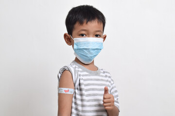 Asian boy wearing face mask giving thumbs up and showing his arm with bandage after getting vaccination. Child immunization. Protect children with vaccine concept. copy space