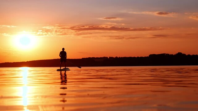 Lonely man stands on the sup board holding an oar. Paddle-boarding on the calm river at sunset. Low angle view.