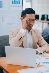 Naklejka premium Excited asian male entrepreneur using smartphone and laptop, working online while sitting at workplace at home office. Businessman texting via cellphone and smiling