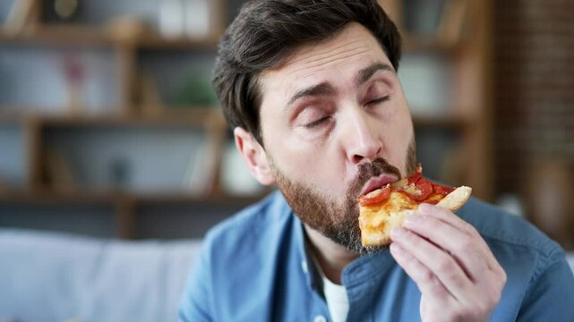 Cheerful satisfied handsome man eating pizza sitting on sofa in living room in apartment. A smiling bearded mature male is happy about the fast delivery of food. Ordering food to take home. Close up