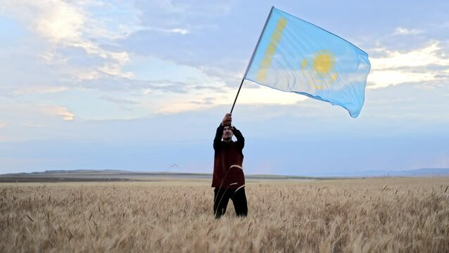 A Kazakh man stands in the steppe of Kazakhstan, holding a dombra. He is dressed in traditional Kazakh clothing, surrounded by tall grass and an expansive sky at sunset.