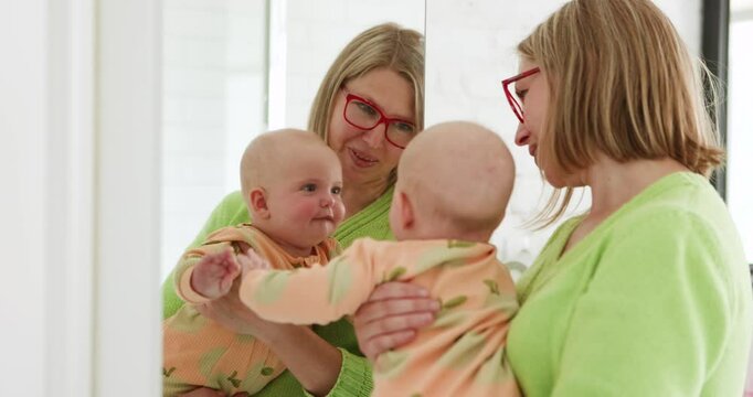 A caring mother in a green sweater is holding her 6 month baby in a yellow outfit with leaf patterns. They are both looking at their reflection in a mirror, showing affection and love for each other
