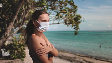 A woman stands on the beach, her face obscure behind a protective mask. Despite the sunny...