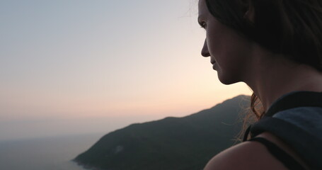 Woman sitting at mountain top, dark dramatic sunset shot. Close up portrait of tourist girl looking on ocean bay and mountain landscape. Travel, tourism, hiking, relax.