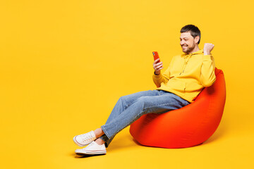 Full body young cool happy man wears hoody casual clothes sit in bag chair hold use mobile cell phone do winner gesture isolated on plain yellow orange background studio portrait. Lifestyle concept.