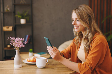 Side view young smart happy student woman wear orange shirt hold use mobile cell phone sitting alone at table in coffee shop cafe restaurant indoors work or study. Freelance office business concept.