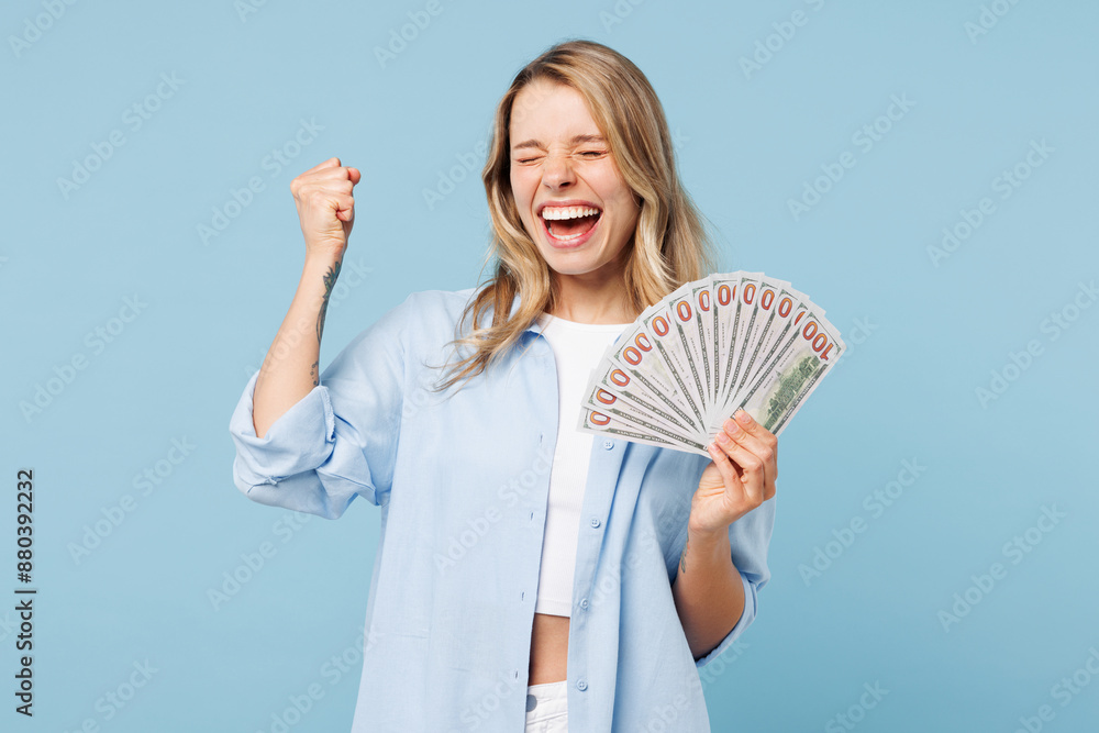 Wall mural Young happy woman she wears white top shirt casual clothes hold in hand fan of cash money in dollar banknotes do winner gesture isolated on plain pastel light blue cyan background. Lifestyle concept.