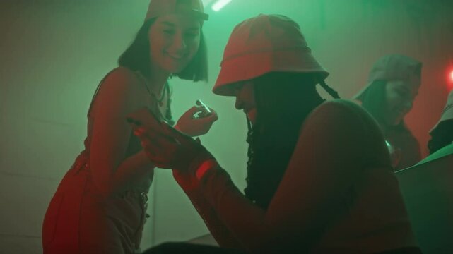 Low angle shot of cheerful African American lady showing pictures of boys on phone to Caucasian girl friend while hanging out in nightclub restroom in y2k outfits