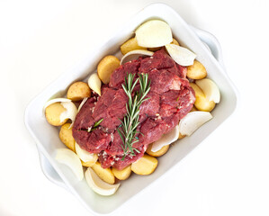 Flat lay of a all beef roast prepared with potatoes, onions and garlic in a white square dish ready for the oven. Isolated on white