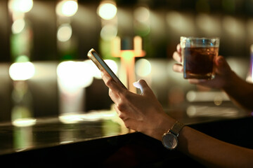 Close up of female hand holding a glass of whisky and using mobile phone at the bar counter