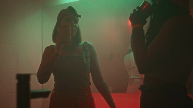 Side shot of fashionable ladies in y2k outfits dancing in smokey nightclub restroom with green and red neon lights and drinking alcohol from disposable cups, hanging out separately