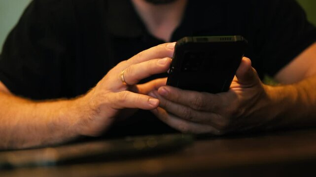 Man uses a phone while sitting at table in modern dining room. Young bearded man at home using cell phone, swiping, tapping and sliding device touch screen. Watching news, reading stories social media