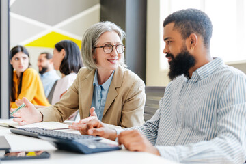 Fototapeta premium Senior businesswoman, leader gesturing while discussing with young male colleague in busy office