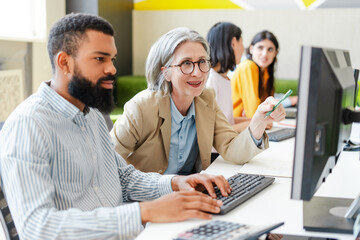 Senior businesswoman, CEO helping young man working on desktop computer in busy office