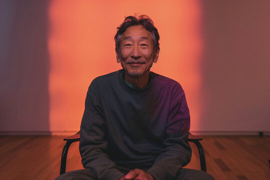 An older man with a calm smile, sitting on a chair against an orange-lit background, exuding warmth, contentment, and relaxed ambiance in a modern indoor setting.