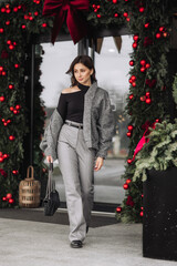 A woman wearing a black jacket and gray pants walks past a Christmas tree. She is carrying a black purse and has a black handbag