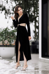 Obraz premium A woman in a black dress with a split skirt is standing in front of a white house. Concept of elegance and sophistication, as the woman is dressed in a formal gown and is posing for the camera