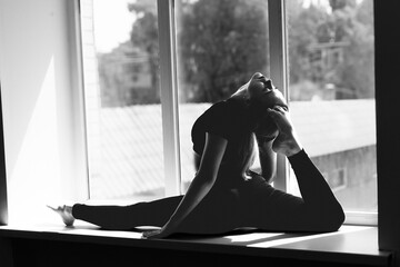 Silhouette of a flexible, slender girl gymnast in the splits against the background of a blurry window. Gymnastics and yoga classes in the studio. Black and white artistic photo