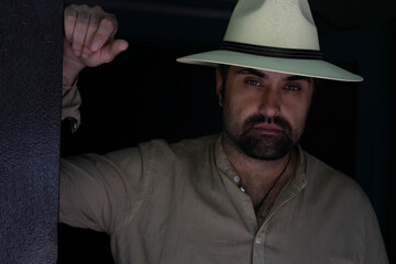 Portrait of a handsome bearded man in a shirt and white hat. A tired man with an expressive gaze looks at the camera, leaning his hand on the door frame. High quality photo