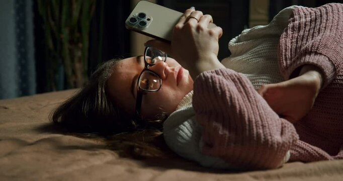 Bored teen girl in glasses using smartphone, wait for important message, interesting information update. Relationship problem. Move phone away with upset. Lying on bed in a sad bad mood