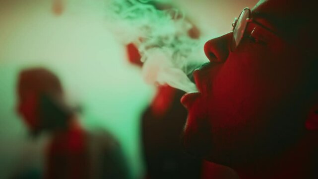 Side closeup of confident young raver in glasses smoking e-cigarette and making smoke rings while hanging out in nightclub with green and red neon lights