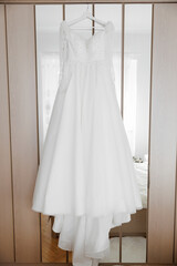 A white wedding dress is hanging on a clothes hanger in a bedroom. The dress is long and has a lot of sparkles on it