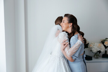 A bride and her bridesmaid are hugging each other. The bride is wearing a white dress and the...