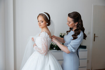 A bride is getting ready for her wedding with the help of her mother. The bride is wearing a long...