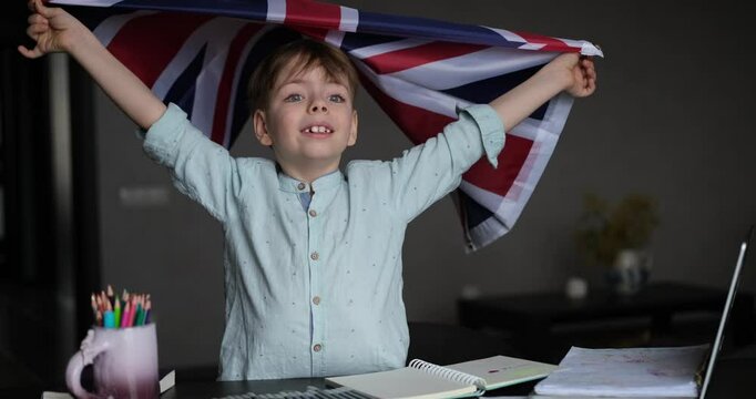 happy boy smiling and holding British flag sitting zt desk with laptop and notes. Concept of English learning at home. video footage 4k