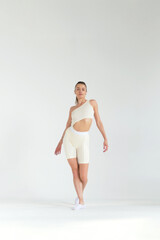 Woman standing in a minimalistic studio, dressed in a beige asymmetric outfit, striking a confident pose. Concept: Emphasizing the blend of fashion and athleticism, highlighting modern fitness trends.