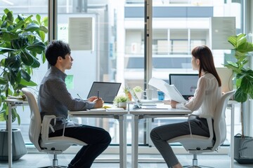 A Japanese man and woman sit at their desks in the office, with white walls behind them. The two people look very serious as they gaze straight ahead during an interview.