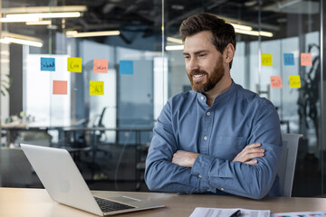 A young smiling young man in a blue shirt and with a beard is sitting in the office at the desk, crossing his arms on his chest and looking at the laptop screen