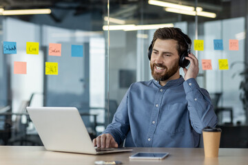 A smiling young man in a headset is working in the office on a laptop, sitting at the workplace and...
