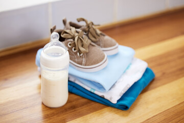 Home, baby clothes and shoes with milk, prepare for toddler and calcium on wooden table. Empty, sneakers and nutrition with fashion for child, dairy and fresh with folded laundry in house with bottle
