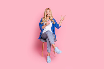 Full size photo of cheerful woman dressed shirt sit on chair hold smartphone directing look empty space isolated on pink color background
