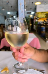 Hand of a man, sitting at a table, holding a glass of white wine, against a restaurant background. Hand of a man with a glass of white wine toasting. Glass of white wine held in hand by a man.