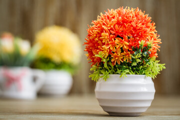 Vibrant, artificial orange,yellow flower arrangements placed on a rustic wooden surface, potted in a white pot that contrasts nicely with the colorful blossoms.